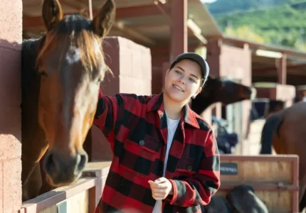 Caring young girl in plaid shirt cleaning and petting purebreed horse with in a countryside club.