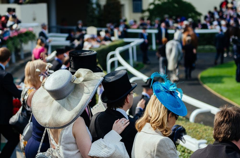 Attendees in formal wear at a horse racing event, with elegant hats and a top hat, conveying a high-profile social gathering.